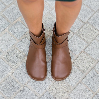A photo of the Shapen Divine ankle boots made with a smooth leather upper, a microline lining, and rubber soles. The boots are brown in color and have an ankle strap with a gold buckle, as well as a side zipper. Both boots are shown from above on a woman’s feet with a view of her knees down. The woman is standing on a gray cobblestone sidewalk. #color_brown