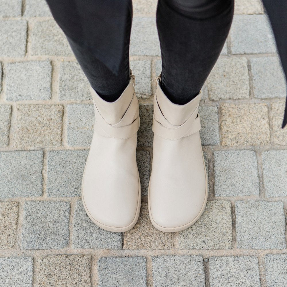 A photo of the Shapen Divine ankle boots made with a smooth leather upper, a microline lining, and rubber soles. The boots are vanilla in color and have an ankle strap with a gold buckle, as well as a side zipper. Both boots are shown from above on a woman’s feet with a view of her shins down. The woman is wearing black skinny jeans tucked into the boots, and she is standing on a gray cobblestone sidewalk. #color_vanilla