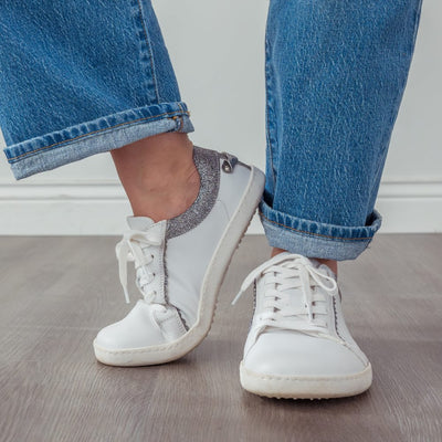 A photo of Shapen Feelin Chic leather sneakers in white. Sneakers have dark grey glitter blocks around the foot opening and a scalopped glitter detail enhancing both sides of the laces. Glitter detail also goes down the back with a glitter pull tab attached by rivets. Both shoes are shown here from the front with the left shoe turned to the side atop a grey wood floor on a womans feet wearing medium wash wide leg blue jeans. #color_white-glitter
