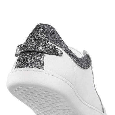A photo of Shapen Feelin Chic leather sneakers in white. Sneakers have dark grey glitter blocks around the foot opening and a scalopped glitter detail enhancing both sides of the laces. Glitter detail also goes down the back with a glitter pull tab attached by rivets. Left shoe is shown from the back here against a white background. #color_white-glitter