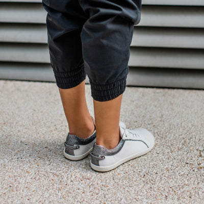 A photo of Shapen Feelin Chic leather sneakers in white. Sneakers have dark grey glitter blocks around the foot opening and a scalopped glitter detail enhancing both sides of the laces. Glitter detail also goes down the back with a glitter pull tab attached by rivets. Both shoes are shown here diagonally from the back right atop pebble pavement on a womans feet wearing dark grey joggers hiked up to the calf. #color_white-glitter