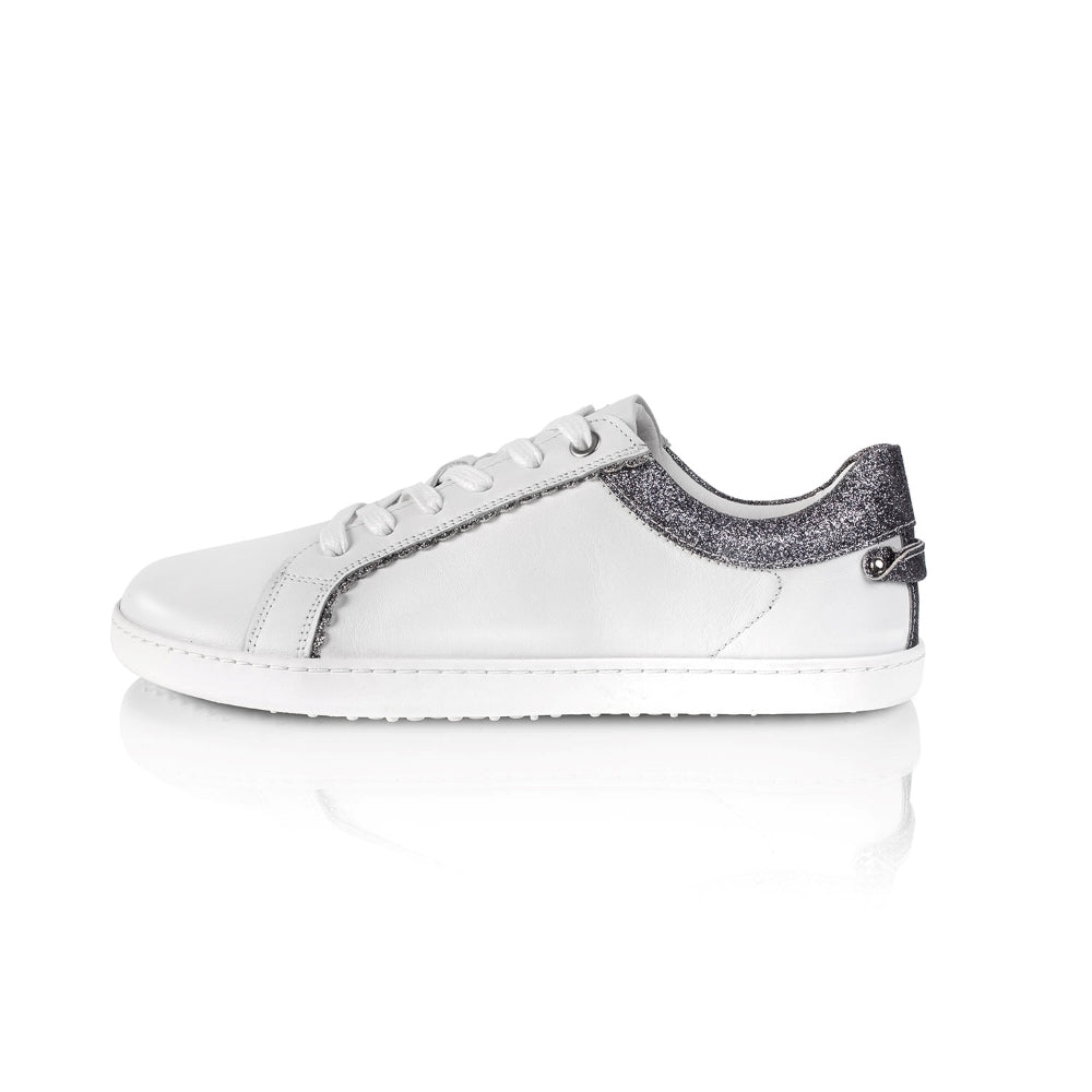 A photo of Shapen Feelin Chic leather sneakers in white. Sneakers have dark grey glitter blocks around the foot opening and a scalopped glitter detail enhancing both sides of the laces. Left shoe is shown facing left here against a white background. #color_white-glitter