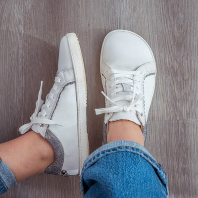 A photo of Shapen Feelin Chic leather sneakers in white. Sneakers have dark grey glitter blocks around the foot opening and a scalopped glitter detail enhancing both sides of the laces. Glitter detail also goes down the back with a glitter pull tab attached by rivets. Both shoes are shown here from the top down with the left shoe tilted on its side against a grey wood floor on a womans feet wearing medium wash wide leg blue jeans. #color_white-glitter