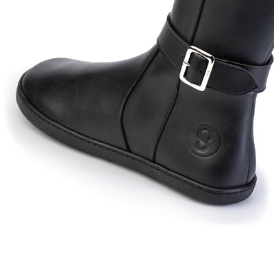 A photo of Shapen Glam lined riding boots made with leather, a faux fur lining, and rubber soles. The boots are black in color with silver buckles on the top and around the ankle, the ankle buckle strap is removable. The left shoe is shown up close from the left side against a white background. #color_black