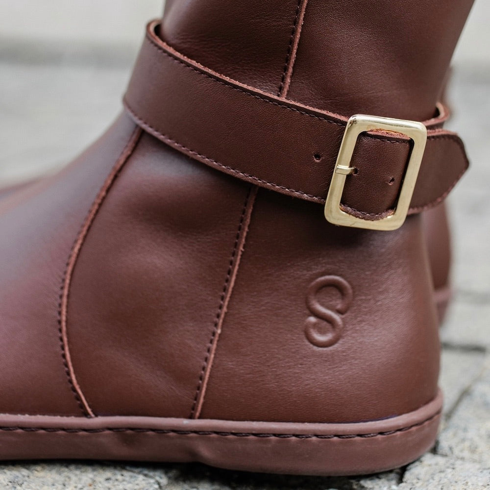 A photo of Shapen Glam lined riding boots made with leather, a faux fur lining, and brown rubber soles. The boots are brown in color with gold buckles on the top and around the ankle, the ankle buckle strap is removable. Left boot ankle buckle detail is shown facing left on a person standing on a grey brick road. #color_brown