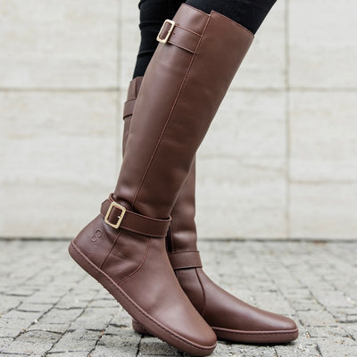 A photo of Shapen Glam lined riding boots made with leather, a faux fur lining, and brown rubber soles. The boots are brown in color with gold buckles on the top and around the ankle, the ankle buckle strap is removable. Both boots are shown facing right on a woman with black skinny jeans tucked in standing on a grey brick road with a grey stone wall in the background. #color_brown