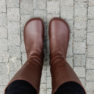 A photo of Shapen Glam lined riding boots made with leather, a faux fur lining, and brown rubber soles. The boots are brown in color with gold buckles on the top and around the ankle, the ankle buckle strap is removable. Both boots are shown from above on a woman with black skinny jeans tucked in standing on a grey brick road. #color_brown