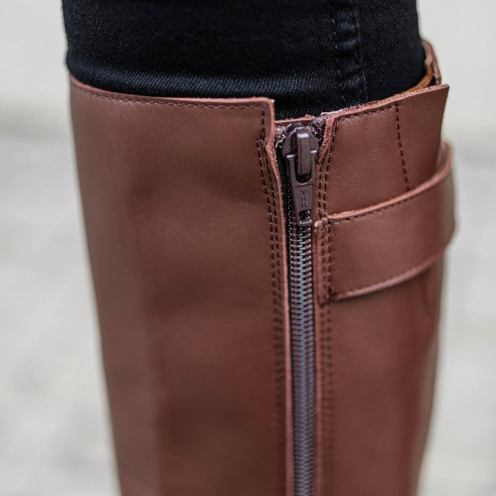 A photo of Shapen Glam lined riding boots made with leather, a faux fur lining, and brown rubber soles. The boots are brown in color with gold buckles on the top and around the ankle, the ankle buckle strap is removable. Right boot zipper detail is shown facing left on a woman with black skinny jeans tucked in standing on a grey brick road with a grey stone wall in the background. #color_brown