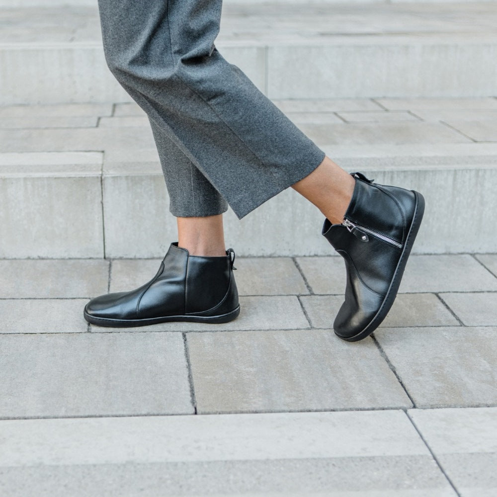 A photo of Shapen Ivy Chelsea boots made from smooth leather and rubber soles. The boots are black in color with stitching detailing and have a silver zipper with a leather tab on the side. Both boots are shown from the left side on a woman’s feet with a view of her knees down. The woman is wearing cropped gray straight leg pants and is walking on pavers, with her right foot in front of her left. #color_black
