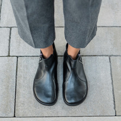 A photo of Shapen Ivy Chelsea boots made from smooth leather and rubber soles. The boots are black in color with stitching detailing and have a silver zipper with a leather tab on the side. Both boots are shown together from above on a woman’s feet, with a view of her knees down. The woman is wearing cropped gray straight leg pants and is standing on gray pavers. #color_black