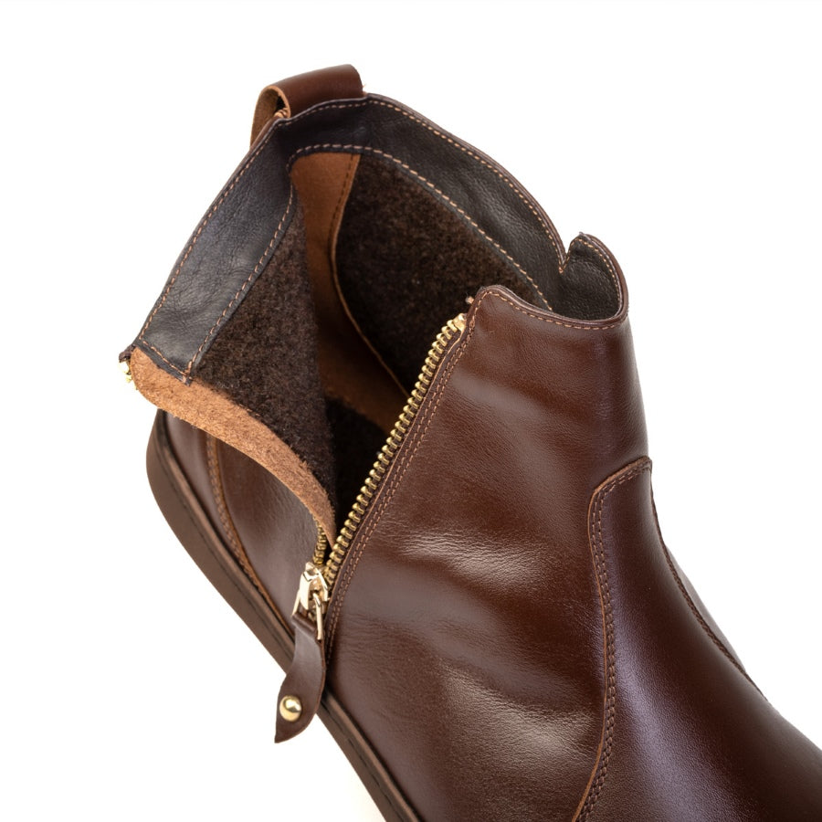 A photo of Shapen Ivy Chelsea boots made from smooth leather and rubber soles. The boots are brown in color with stitching detailing and have a gold zipper with a leather tab on the side. One boot is shown unzipped to to show the fleece inside against a white background. #color_brown