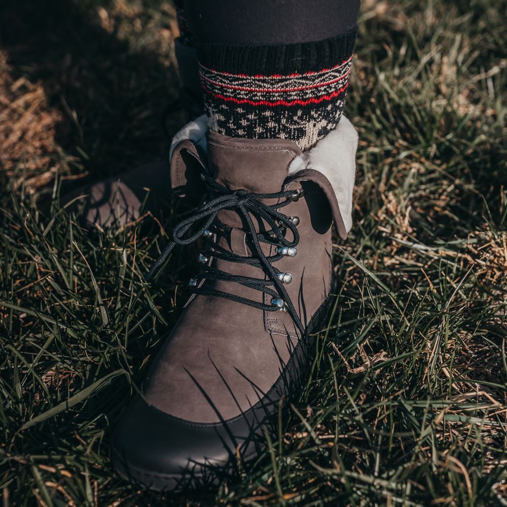 A photo of the Shapen Lynx lace up boots made from a water-resistant nubuck leather uppers and black rubber soles. The boots are dark grey in color, have a black leather rim around the bottom inch of the shoe, black laces, and a white wool lining that can be shown by folding over the top of the boots. The left boot is shown on a woman's foot from the front, with a view of her shins down. The woman is wearing black leggings tucked into holiday themed socks and is standing on grass. #color_dark-grey