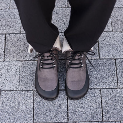 A photo of the Shapen Lynx lace up boots made from a water-resistant nubuck leather uppers and black rubber soles. The boots are dark grey in color, have a black leather rim around the bottom inch of the shoe, black laces, and a white wool lining that can be shown by folding over the top of the boots. Both boots are shown on a man's feet from above with a view of his knees down. The man is wearing black pants and is standing on a paved sidewalk. #color_dark-grey