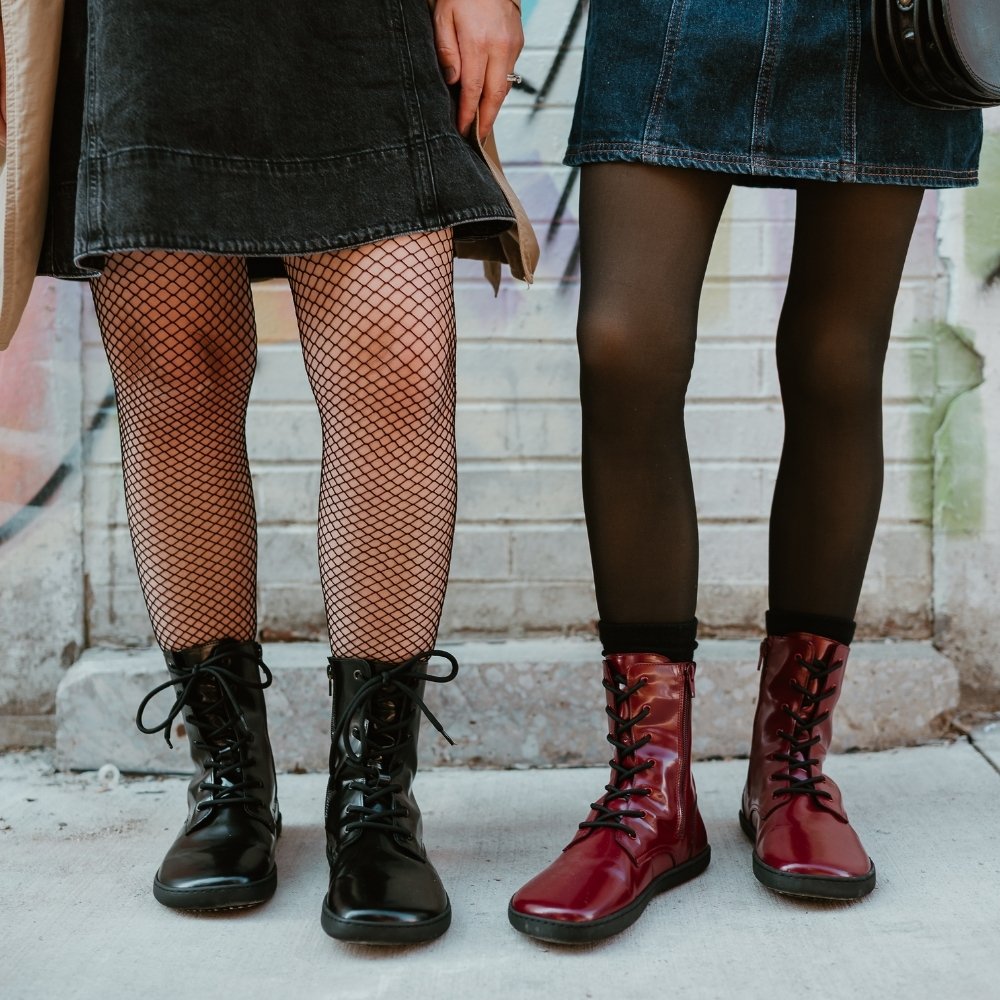Dark red leather Shapen Maverick combat boots with a red, branded pull tab, a side zipper, black laces, and black rubber soles. Both shoes are shown from the front on a light-skinned woman wearing a blue-jean jumper with sheer black tights, and a black leather purse standing on a sidewalk in an urban setting to the right of another light-skinned woman wearing the black maverick boot. #color_bordeaux