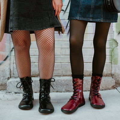 Dark red leather Shapen Maverick combat boots with a red, branded pull tab, a side zipper, black laces, and black rubber soles. Both shoes are shown from the front on a light-skinned woman wearing a blue-jean jumper with sheer black tights, and a black leather purse standing on a sidewalk in an urban setting to the right of another light-skinned woman wearing the black maverick boot. #color_bordeaux