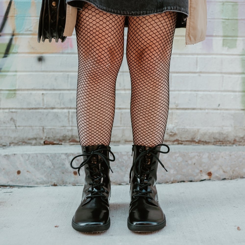 Black leather Shapen Maverick combat boots with a black, branded pull tab, a side zipper, black laces, and black rubber soles. Both shoes are shown from the front on a light-skinned woman  standing on a sidewalk in an urban setting wearing fishnet tights, a dark-grey jean jumper, tan trench coat, and a brown leather purse. #color_black