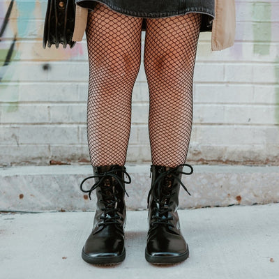 Black leather Shapen Maverick combat boots with a black, branded pull tab, a side zipper, black laces, and black rubber soles. Both shoes are shown from the front on a light-skinned woman  standing on a sidewalk in an urban setting wearing fishnet tights, a dark-grey jean jumper, tan trench coat, and a brown leather purse. #color_black