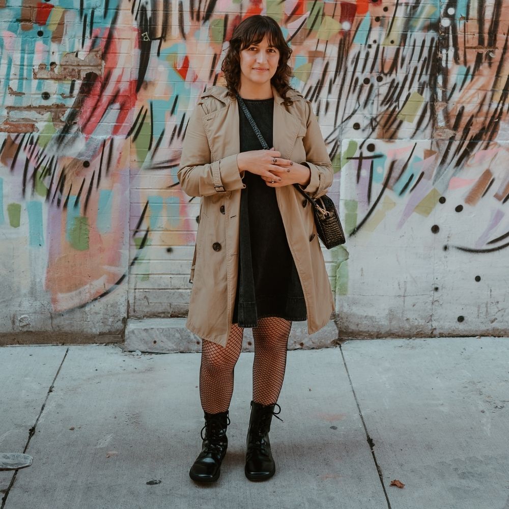 Black leather Shapen Maverick combat boots with a black, branded pull tab, a side zipper, black laces, and black rubber soles. Both shoes are shown from the front on a light-skinned woman, with short, curly, brown hair  standing on a sidewalk in an urban setting wearing fishnet tights, a dark-grey jean jumper, tan trench coat, and a brown leather purse. #color_black