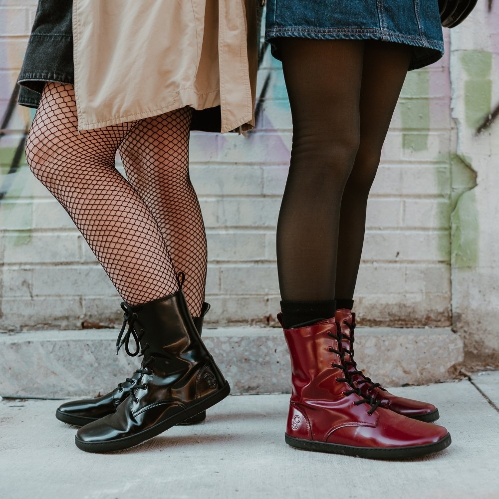 Black leather Shapen Maverick combat boots with a black, branded pull tab, a side zipper, black laces, and black rubber soles. Both shoes are shown facing left on a light-skinned woman  standing on a sidewalk in an urban setting wearing fishnet tights, a dark-grey jean jumper, tan trench coat, and a brown leather purse standing next to another light-skinned woman wearing the Bordeaux Maverick boots. #color_black
