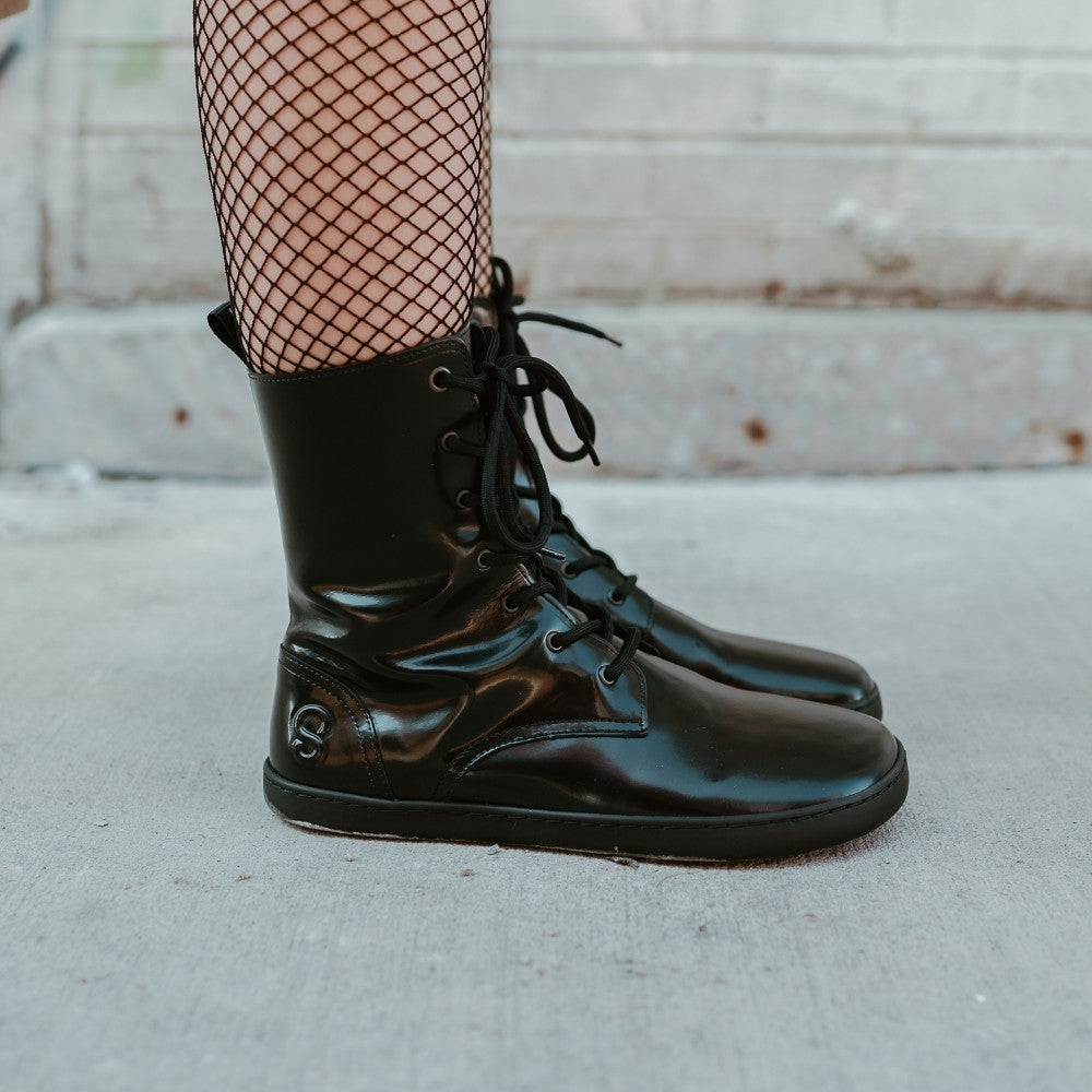 Black leather Shapen Maverick combat boots with a black, branded pull tab, a side zipper, black laces, and black rubber soles. Both shoes are shown facing right on a light-skinned woman  standing on a sidewalk in an urban setting wearing fishnet tights. #color_black