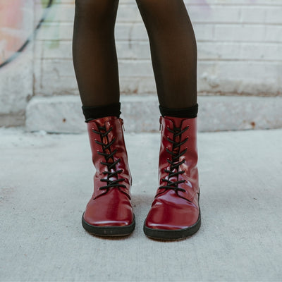 Dark red leather Shapen Maverick combat boots with a red, branded pull tab, a side zipper, black laces, and black rubber soles. Both shoes are shown from the front on a light-skinned woman wearing sheer black tights standing on a sidewalk in an urban setting. #color_bordeaux