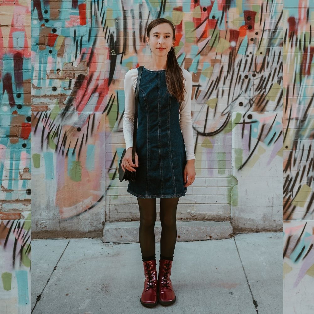 Dark red leather Shapen Maverick combat boots with a red, branded pull tab, a side zipper, black laces, and black rubber soles. Both shoes are shown from the front on a light-skinned woman with long, straight, brown hair wearing a blue-jean jumped with a white long-sleeve shirt, sheer black tights, and a black leather purse standing on a sidewalk in an urban setting. #color_bordeaux