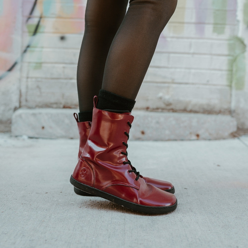 Dark red leather Shapen Maverick combat boots with a red, branded pull tab, a side zipper, black laces, and black rubber soles. Both shoes are shown facing right on a light-skinned woman wearing sheer black tights standing on a sidewalk in an urban setting. #color_bordeaux