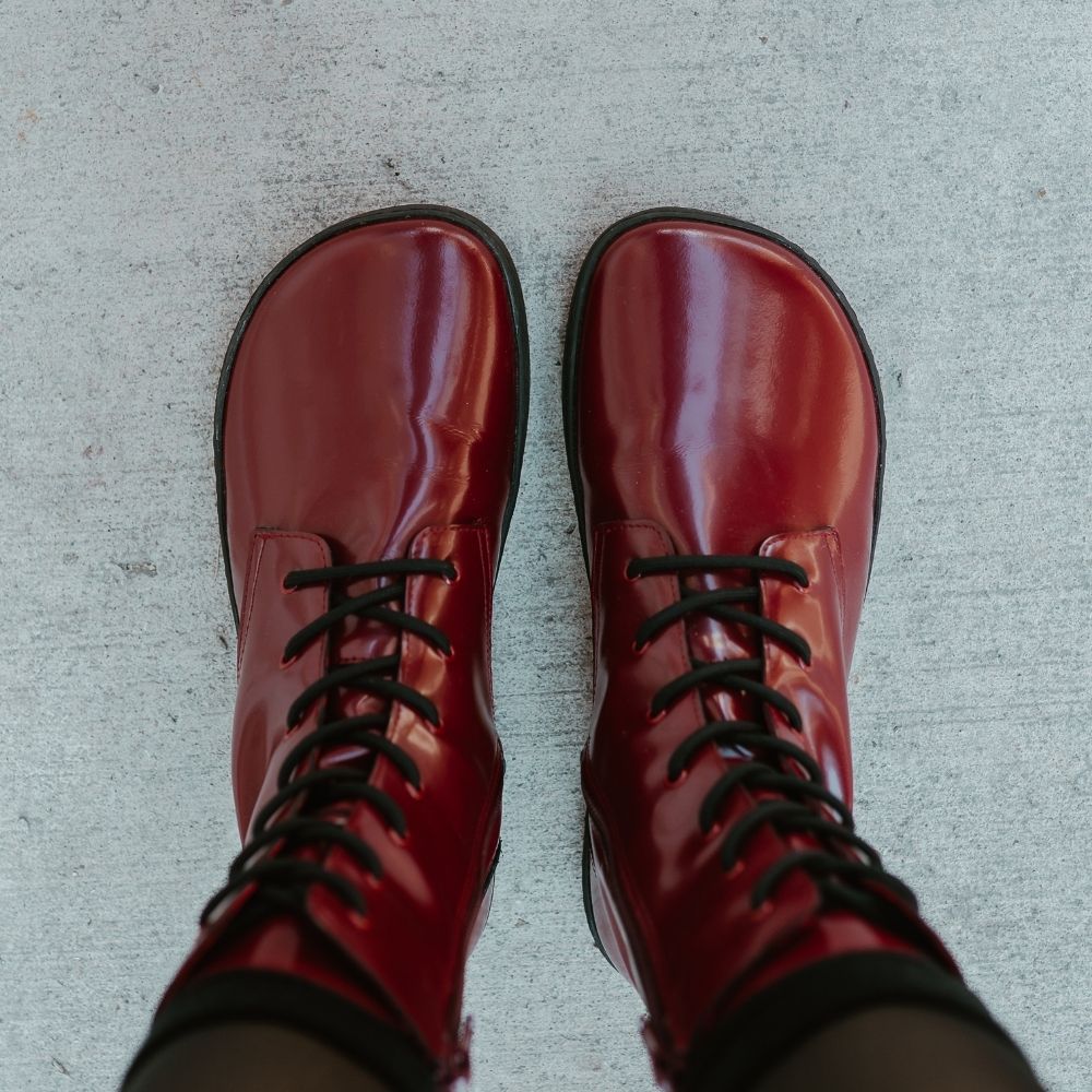 Dark red leather Shapen Maverick combat boots with a red, branded pull tab, a side zipper, black laces, and black rubber soles. Both shoes are shown from above on a light-skinned woman wearing sheer black tights standing on a sidewalk in an urban setting. #color_bordeaux