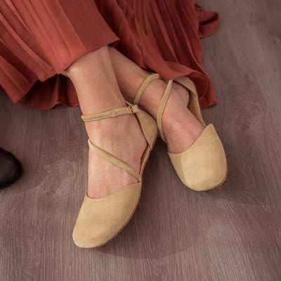 A photo of Beige Shapen Orchid flats.  Shoes are suede with tan rubber soles. A toe box and heel cup bookmark each end of the shoe with a round strap that zig zags from the toe box to the ankle/heel cup to keep the shoes secured to the foot. Both shoes are shown here from above facing slightly left on a woman with light skin sitting on a grey wood floor wearing a burnt orange pleated dress. #color_beige