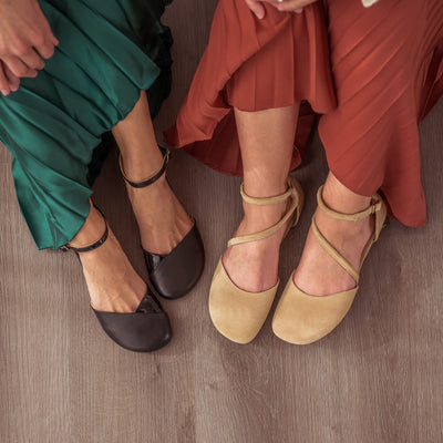 A photo of Beige Shapen Orchid flats.  Shoes are suede with tan rubber soles. A toe box and heel cup bookmark each end of the shoe with a round strap that zig zags from the toe box to the ankle/heel cup to keep the shoes secured to the foot. Both shoes are shown here from above facing slightly left on a woman with light skin sitting on a grey wood floor wearing a burnt orange pleated dress. To the left of her is another woman wearing black Shapen Poppy flats wearing a green silky pleated dress. #color_beige
