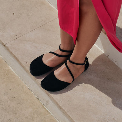 A photo of black Shapen Orchid flats. Shoes are suede with tan rubber soles. A toe box and heel cup bookmark each end of the shoe with a round strap that zig zags from the toe box to the ankle/heel cup to keep the shoes secured to the foot. Both shoes are shown here from the top on a dark-skinned woman wearing a min-length silky pink dress sranding on smooth tan stairs. #color_black
