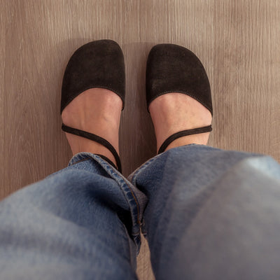 A photo of black Shapen Orchid flats. Shoes are suede with tan rubber soles. A toe box and heel cup bookmark each end of the shoe with a round strap that zig zags from the toe box to the ankle/heel cup to keep the shoes secured to the foot. Both shoes are shown here from above on a woman wearing light-wash, wide-legged jeans standing on a grey wooden floor. #color_black