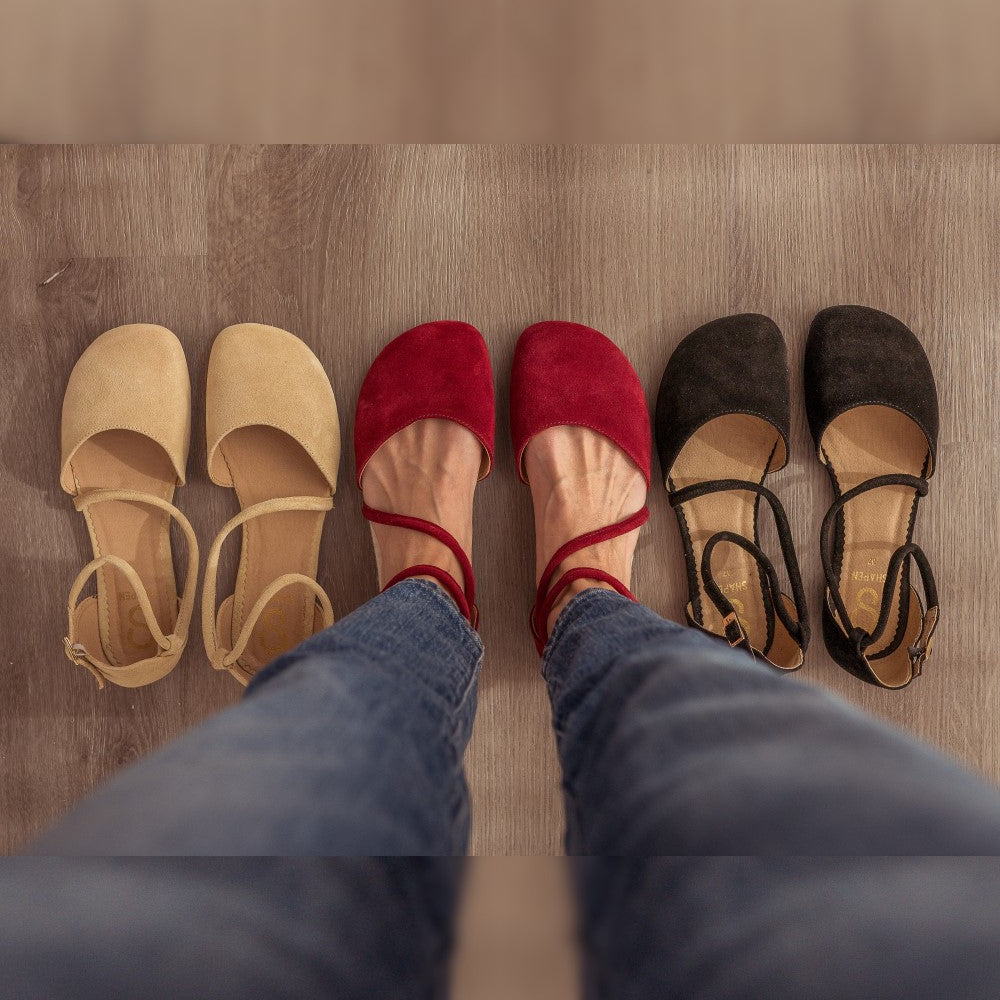A photo of dark red Shapen Orchid flats. Shoes are suede with tan rubber soles. A toe box and heel cup bookmark each end of the shoe with a round strap that zig zags from the toe box to the ankle/heel cup to keep the shoes secured to the foot. Both shoes are shown here from above  on a woman wearing medium-wash skinny jeans standing on a grey wooden floor. Beige Orchid flats are to the left and black orchid flats are to the right.  #color_bordeaux