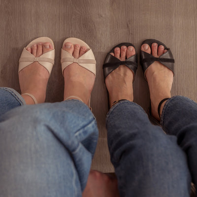 Light Rose Shapen Petal Sandals. Sandals are a peep toe style at the toe box and a heel cup attached to a thin, suede ankle strap. Both shoes are shown here from above on a woman wearing light-wash, wide-legged jeans sitting on a bench on a grey wooden floor with a woman wearing black Petal flats and medium-wash skinny jeans to the right. #color_light-rose