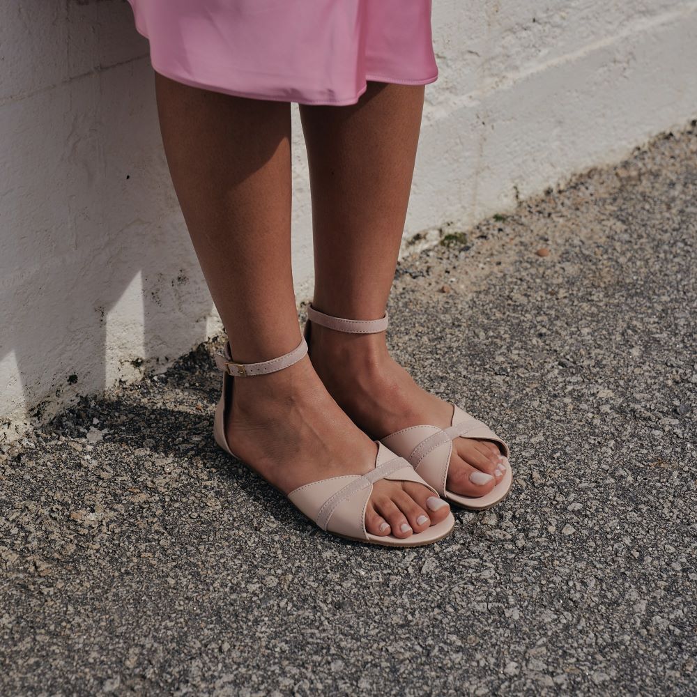 Light Rose Shapen Petal Sandals. Sandals are a peep toe style at the toe box and a heel cup attached to a thin, suede ankle strap. Both shoes are shown diagonally to the right on a dark-skinned woman with short, black, curly hair wearing a silk, pink, mid-lenth skirt standing on grey pavement against a white brick wall. #color_light-rose