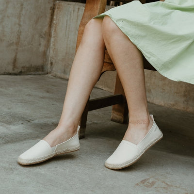 Beige canvas Shapen Espadrilles loafers with a pull tab and tan soles. Both shoes are shown facing diagonally left on a light-skinned woman wearing a knee-length seafoam green dress sitting on a wooden bench with cement walls and floors in the background. #color_beige