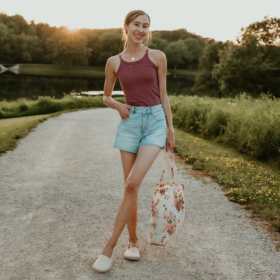Beige canvas Shapen Espadrilles loafers with a pull tab and tan soles. Both shoes are shown on a light-skinned woman with long brown hair wearing a brick red tank top tucked into light-wash jean shorts, carrying a floral bag on a path in nature. #color_beige