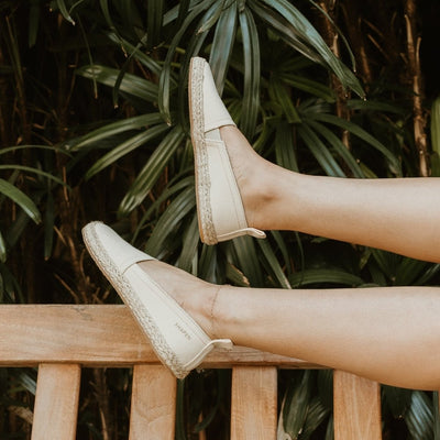 Beige canvas Shapen Espadrilles loafers with a pull tab and tan soles. Both shoes are shown facing upwards on a light-skinned woman leaning back on a wooden bench with foliage in the background. #color_beige
