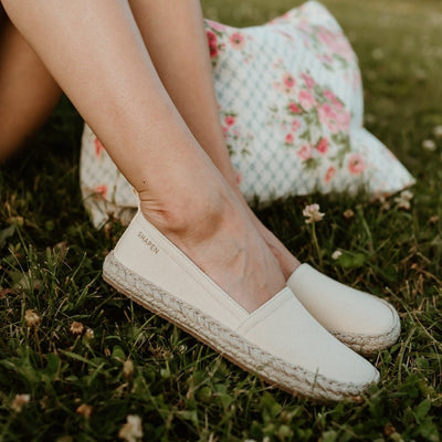 Beige canvas Shapen Espadrilles loafers with a pull tab and tan soles. Both shoes are shown on facing right a light-skinned woman sitting in grass with a floral bag behind. #color_beige