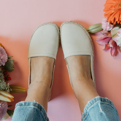 Beige canvas Shapen Espadrilles loafers with a pull tab and tan soles. Both shoes are shown from above on a tan woman wearing ankle length light wash jeans sitting on a pink floor with colorful flowers surrounding. #color_beige