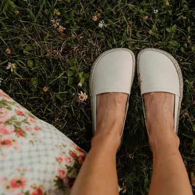 Beige canvas Shapen Espadrilles loafers with a pull tab and tan soles. Both shoes are shown from above a light-skinned woman sitting in grass with a floral bag to the left. #color_beige
