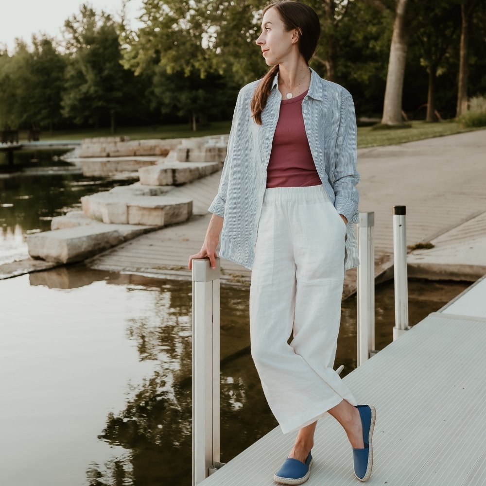 Indigo canvas Shapen Espadrilles loafers with a pull tab and tan soles. Both shoes are shown on a light-skinned woman with long brown hair wearing white linen pants, a red shirt, and a long-sleeve blue linen button up standing on a metal dock by the water. #color_indigo