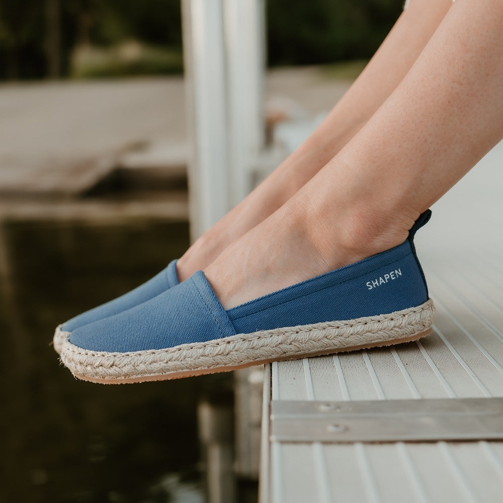 Indigo canvas Shapen Espadrilles loafers with a pull tab and tan soles. Both shoes are shown facing left on a light-skinned woman sitting on a metal dock by the water. #color_indigo