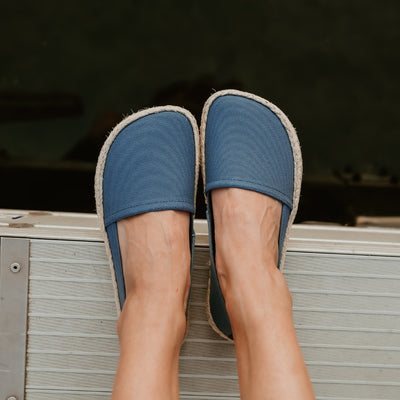 Indigo canvas Shapen Espadrilles loafers with a pull tab and tan soles. Both shoes are shown from above on a light-skinned woman sitting on a metal dock by the water. #color_indigo