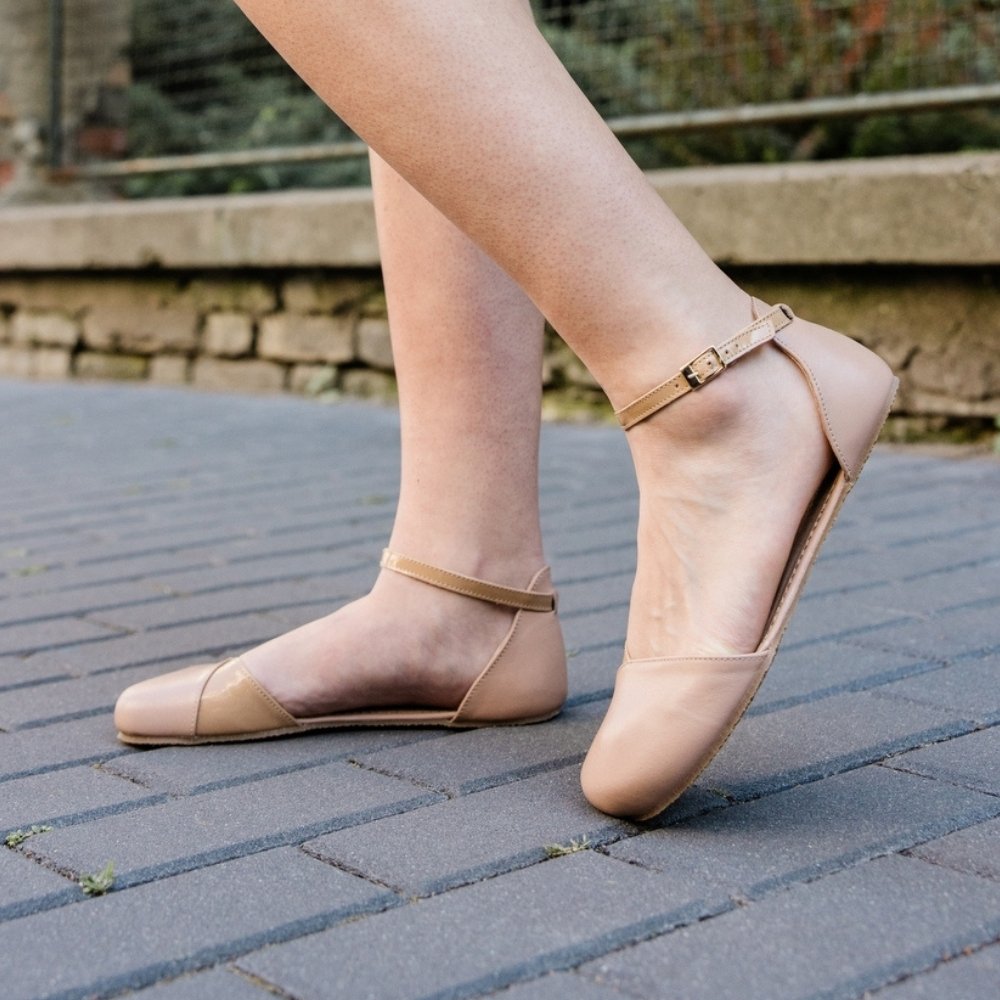 Beige, leather Shapen Poppy D’orsay dressy flats with tan rubber soles. The flats have a heel cup and dainty patent leather ankle straps. Both shoes are shown facing left on a light-skinned woman with the left foot propped up on the toe behind the right on a grey brick pathway with a stone wall and fence in the background. #color_beige 