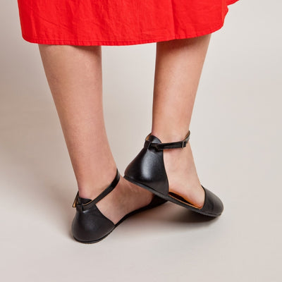 A photo of Shapen Poppy D’orsay dressy flats with a leather upper and rubber soles. The flats are a black color with a heel cup and dainty ankle strap, the inside of the sole are a beige color. Both flats are shown diagonally from the back right against a cream background on a woman with long brown hair wearing a mid-length bright red button down dress. Her right heel is lifted. #color_black