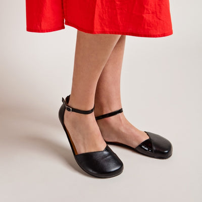 A photo of Shapen Poppy D’orsay dressy flats with a leather upper and rubber soles. The flats are a black color with a heel cup and dainty ankle strap, the inside of the sole are a beige color. Both flats are shown diagonally from the front right against a cream background on a woman with long brown hair wearing a mid-length bright red button down dress. Her right heel is lifted. #color_black