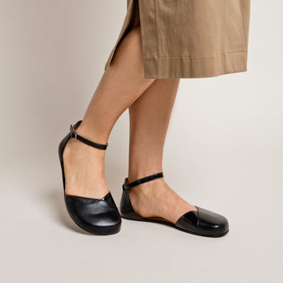 A photo of Shapen Poppy D’orsay dressy flats with a leather upper and rubber soles. The flats are a black color with a heel cup and dainty ankle strap, the inside of the sole are a beige color. Both flats are shown from the front against a cream background on a woman wearing a mid-length tan button down. #color_black