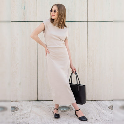 Black leather Shapen Poppy D’orsay dressy flats with a tan footbed and tan rubber soles. The flats have a heel cup held on by a dainty, patent leather ankle strap. Both shoes are shown from the front on a light-skinned woman with short, light-brown hair. She is standing on a stone tile pathway wearing a long beige dress and tan sunglasses holding a black leather bag with her left foot crossed over the right and left hand on her hip. #color_black