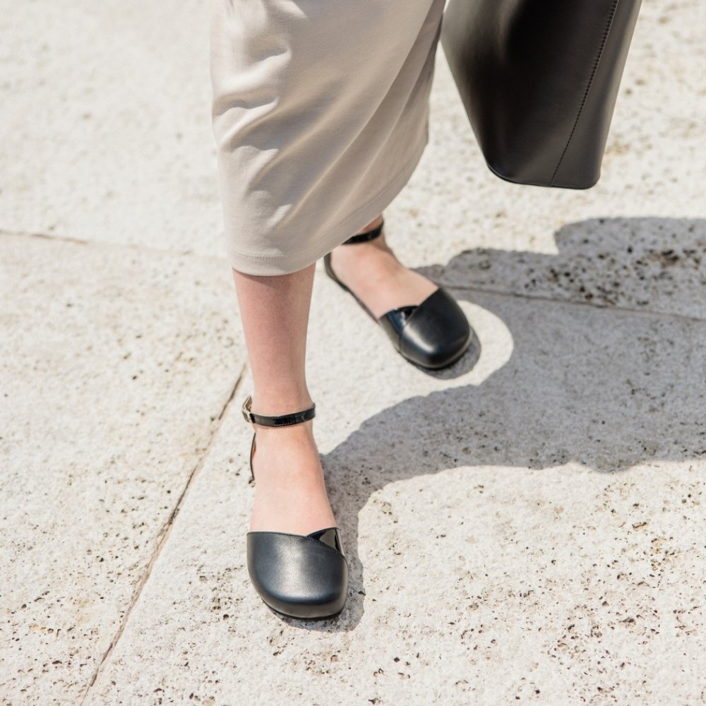 Black leather Shapen Poppy D’orsay dressy flats with a tan footbed and tan rubber soles. The flats have a heel cup held on by a dainty, patent leather ankle strap. Both shoes are shown staggered slightly above from the front on a light-skinned woman. She is standing on a stone tile pathway wearing a long beige dress holding a black leather purse on the right. #color_black